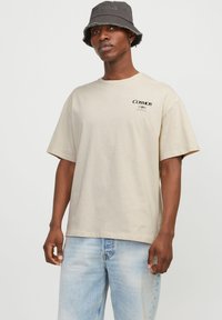 T-shirt en coton beige à manches courtes, encolure ronde et logo "COSMOS" noir sur la poitrine. Assorti à un jean en denim bleu clair.