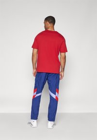 Červené bavlněné tričko s krátkým rukávem; modré sportovní kalhoty s červenými a bílými diagonálními pruhy; nošeno s bílými teniskami. Jednoduché pozadí.