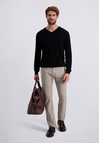 Schwarzer Langarm-V-Ausschnitt-Pullover, beigefarbene Hose, braune Ledertasche und braune Schuhe. Das Model steht vor einem schlichten Hintergrund.