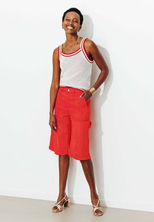 Femme souriante aux cheveux courts, portant un débardeur en maille blanche avec un bord rouge, un short rouge vif arrivant aux genoux et des sandales beiges à talons.