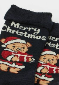 Marineblaue Socken mit einem gestrickten Motiv eines Bären in einem Weihnachtsmann-Outfit, der ein Geschenk hält. Der Text lautet "Frohe Weihnachten" in Weiß. Weiche Textur.