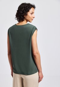 Maglietta verde scuro a maniche corte con scollatura rotonda e una texture liscia. Presenta linee pulite e una vestibilità rilassata, abbinata a pantaloni di colore chiaro.