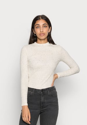 Top de manga longa, de cor creme, com uma textura canelada e padrão salpicado. Decote alto e design ajustado. Combinado com jeans escuros.