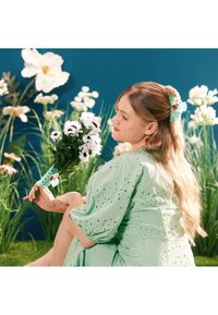 Femme en robe verte tenant des marguerites blanches, assise devant des fleurs hautes avec un fond bleu, cheveux partiellement attachés avec une pince assortie.