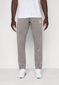 Lichtgrijze joggerbroek gemaakt van zachte stof, met elastische boorden en een wit logo op de linker bovenbeen. Draagt met witte sportschoenen.