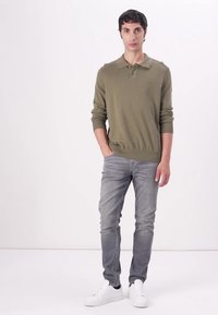 Olivgrüner gestrickter Pullover mit einem Kragen, kombiniert mit hellgrauen Jeans und weißen Sneakern. Glatte Textur, figurbetontes Design.