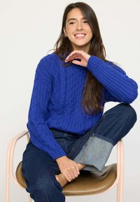 Pull en maille bleu avec motif en cable, manches longues et poignets côtelés. Modèle portant un jean foncé avec le revers replié, assis sur une chaise.
