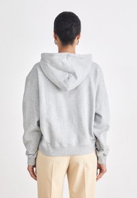 Hoodie supradimensionat gri, cu șnur, manșete și tiv cu zale. Purtați cu pantaloni bej, văzut din spate, arătând gluga și croiala lejeră.