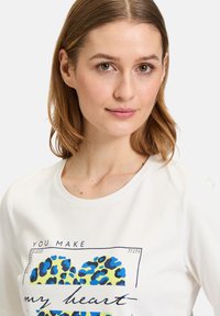 Chemise blanche à manches courtes ornée d'un imprimé graphique représentant un cœur léopard bleu et jaune, avec du texte marine au-dessus et des accents de strass.