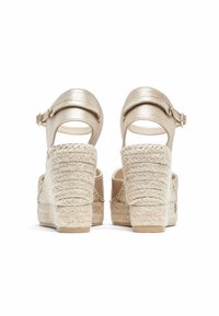 Sandales compensées en espadrille beiges avec des brides métalliques à la cheville, une plateforme en jute texturé et une base de talon solide. Coutures détaillées visibles.