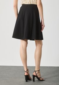 Black A-line skirt with pleats, made from textured fabric. Features a mid-rise waistband. Worn with black heeled sandals.