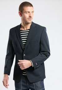 Veste bleu marine avec fermeture à boutons sur le devant, une poche poitrine unique et une chemise à rayures contrastées en dessous. Portée avec un jean foncé, mettant en valeur des lignes épurées.