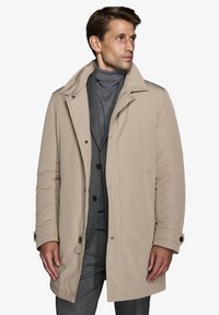 Beige polstret jakke med høj krave, der har en frontlukning, to sidelommer og knapdetaljer på ærmerne. Glat tekstur.