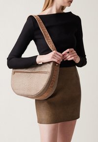 Beige strukturierte Schultertasche mit geschwungener Silhouette, ausgestattet mit einer Reißverschlusstasche und einem markierten Schulterriemen in kontrastierender Farbe.