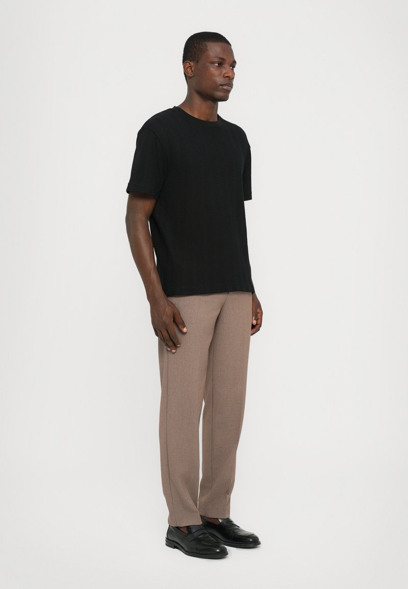 Black knit short-sleeve t-shirt paired with beige tailored trousers and black loafers. Model stands against a plain white background.