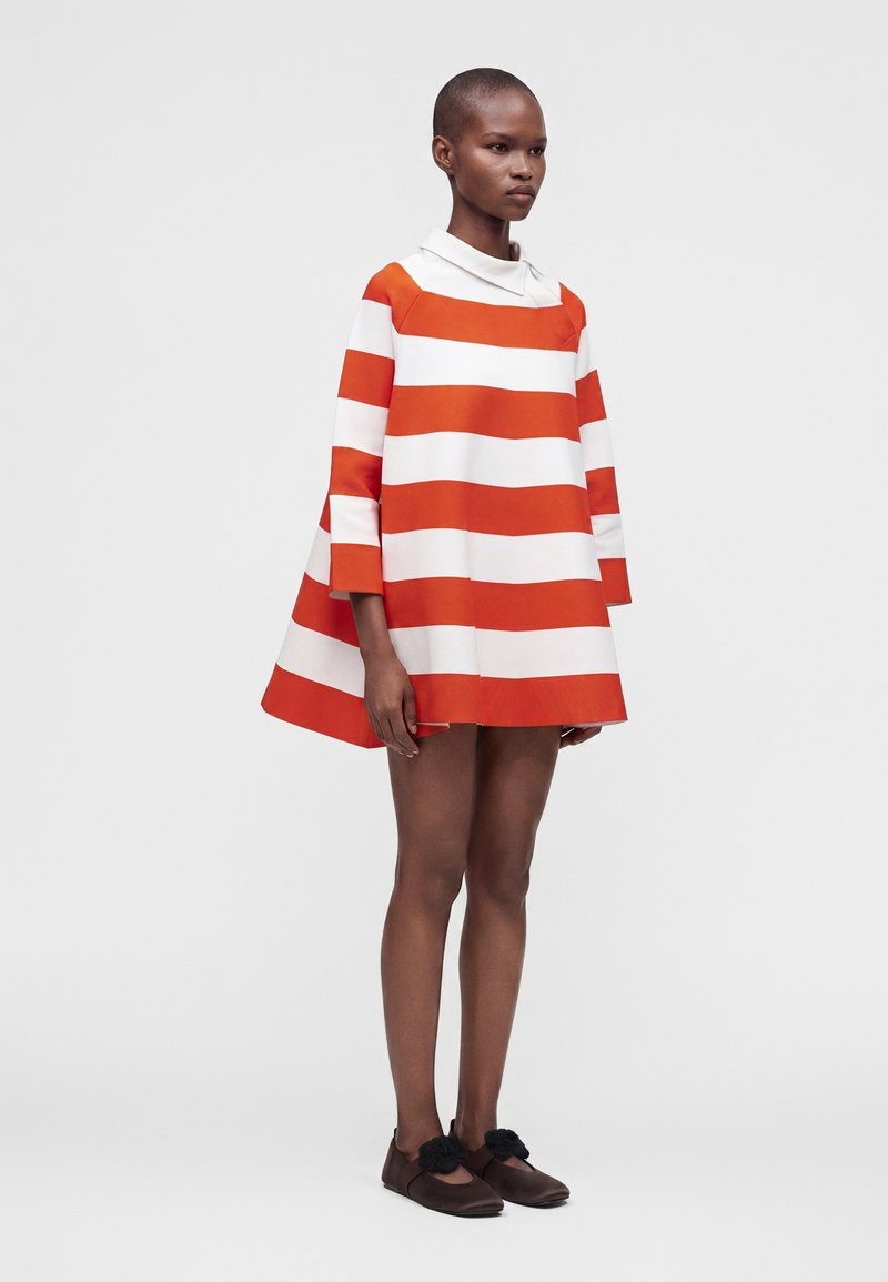 Mannequin portant une robe courte et ample à rayures horizontales orange et blanches et des chaussures marron avec pompons noirs, debout sur un fond uni.