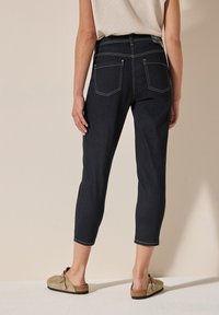 Los jeans recortados de mezclilla oscura presentan un pespunte en contraste, una silueta ajustada y dos bolsillos traseros. Combinados con una blusa clara y zapatos sin cordones.