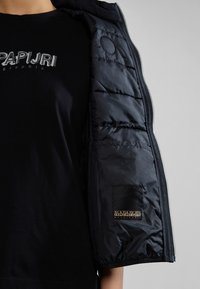 Svart pufferjacka med en glänsande yta, med dragkedja, elastiska ärmmuddar och en logotyp på vänster ärm.