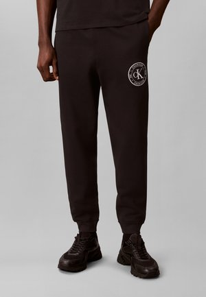 Άτομο που φοράει μαύρη φόρμα Calvin Klein με λογότυπο στον μηρό και μαύρα αθλητικά, στέκεται μπροστά σε μονόχρωμο γκρι φόντο.