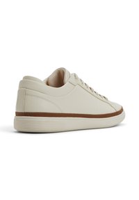 Zapatilla color crema con parte superior de cuero texturizado, forro suave, media suela de goma marrón y suela exterior de goma blanca delgada. Diseño minimalista.