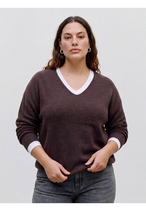 Femme aux cheveux bruns ondulés portant un pull marron col en V avec une bordure blanche et un jean gris, debout devant un fond clair uni.