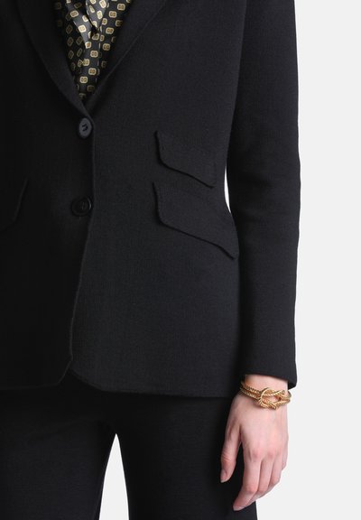 Personne portant un blazer noir avec des poches à rabat, une chemise à motifs et un bracelet en corde torsadée dorée au poignet droit.