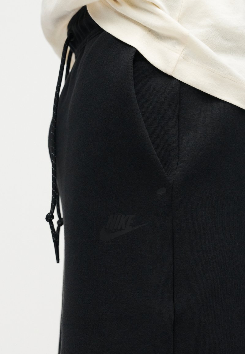 Gros plan sur un pantalon de survêtement noir Nike avec taille à cordon et logo discret près de la poche, assorti à un haut de couleur claire.