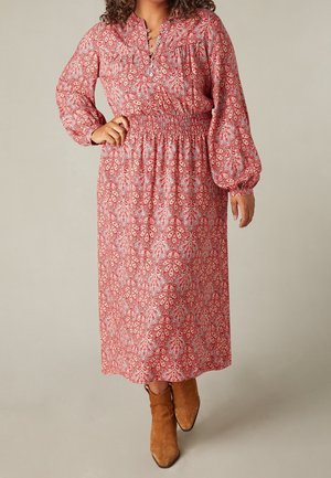 Donna che indossa un vestito rosso a maniche lunghe con fantasia, vita arricciata e stivaletti in camoscio beige, in piedi davanti a uno sfondo uniforme.