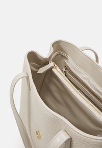 Světlý béžový tote bag s texturovaným vnějším povrchem, dvěma vnitřními přihrádkami, jednou zapínací kapsou a zlatými kovovými akcenty.