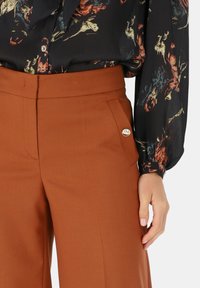 Talia i ręka osoby ubranej w brązowe spodnie z boczną kieszenią oraz czarną bluzkę w wielokolorowy kwiatowy wzór.