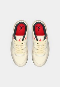 Ανοιχτόχρωμα αθλητικά παπούτσια με υφασμάτινη υφή, κόκκινες λεπτομέρειες, μαύρα στοιχεία και μια επιβλητική λογότυπο στη γλώσσα. Οι κορδόνια είναι μπεζ.