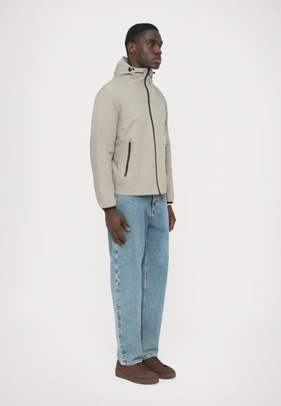 Helebeež kapuutsiga jakk tekstuurse kangaga, must lukuga ja küljetaskutega, kombineeritud helesiniste teksade ja pruunide tossudega.