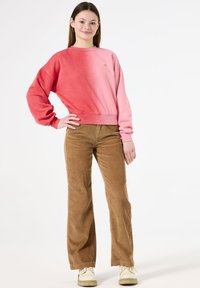 Felpa con maniche lunghe in gradiente rosa e rosso, abbinata a pantaloni in corda a gamba larga di colore marrone. Le scarpe sono chiare e con i lacci.