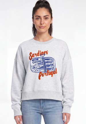 Jeune femme aux cheveux noirs en queue de cheval portant un sweat-shirt gris clair avec une boîte de sardines bleue et un texte orange "Sardines Portugal".
