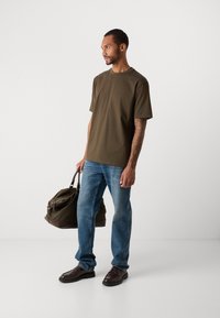 Bărbat cu brațul tatuat, îmbrăcat cu un tricou verde măsliniu, blugi albaștri și pantofi maro, ținând o geantă de voiaj din piele verde închis și maroniu.
