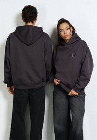 Dwie osoby w ciemnych bluzach z kapturem i czarnych jeansach stoją przy białej ścianie, jedna zwrócona twarzą do przodu, druga plecami.