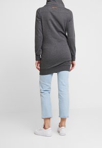 Szara tunika z długim rękawem i wysokim kołnierzem, z tylną metką, zestawiona z jasnoniebieskimi dżinsami i białymi trampkami. Gładka faktura.