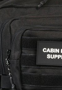 Mochila táctica negra que presenta múltiples compartimentos con cremallera, una superficie texturizada y un parche de PVC etiquetado como "CABIN SUPPLY". Incluye sistema de molle.