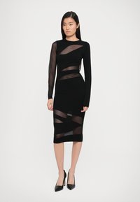 Vestito bodycon nero con maniche lunghe in rete, caratterizzato da ritagli geometrici trasparenti. L'orlo è sotto il ginocchio, abbinato a tacchi alti neri.