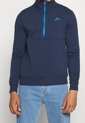 Mann trägt dunkelblauen Nike-Pullover mit blauem Viertel-Reißverschluss und hellblauen Jeans, steht vor einem einfarbigen Hintergrund.