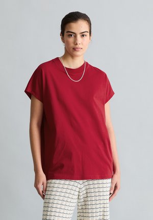 Vrouw die een loszittend rood shirt met korte mouwen, een zilveren ketting en witte broek met patroon draagt, staat voor een egale grijze achtergrond.