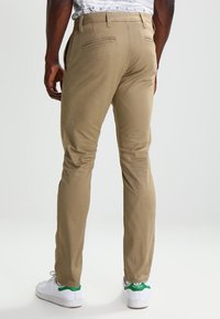 Chinos khaki con vestibilità slim, caratterizzati da una trama di tessuto liscia, doppie tasche posteriori e passanti per la cintura, abbinati a sneakers bianche.