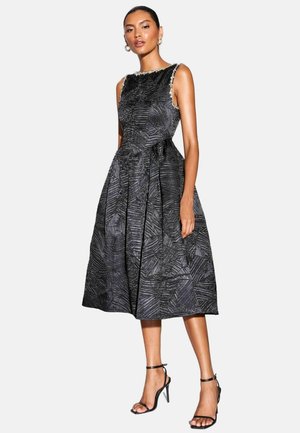 Schwarzes ärmelloses Kleid mit strukturiertem Muster, ausgestelltem Rock und verzierten Halsabschlüssen. Modell trägt schwarze Riemchenheels.
