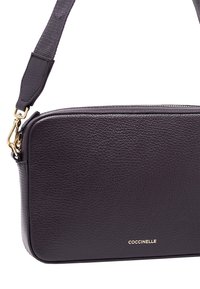 Leren crossbody tas in diep paars met een geutterde textuur, voorzien van een gouden rits en het merkembleem aan de voorkant. Verstelbare schouderband.