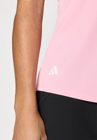 Svetloružový športový top vyrobený z priedušného materiálu, s zakriveným lemom a bielym logom umiestneným v dolnom ľavom rohu. Nosí sa s tmavými legínami.