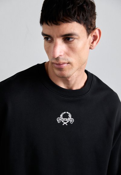 Svart sweatshirt med en texturerad rund hals och en vit broderad logotyp med en dekorativ design på framsidan.