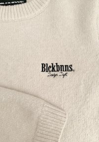 Beige gebreide trui met een ronde halslijn en ribgebreide manchetten, met een zwart geborduurd logo "Blckbnns Design Dept" op de voorkant.