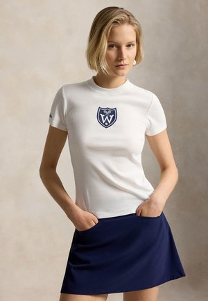 Kvinde iført en hvid figursyet T-shirt med et blåt emblem og et mørkeblåt skørt, hænderne i lommerne, stående foran en ensfarvet beige baggrund.