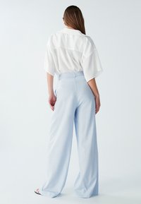 Svetlo modri široke hlače iz mehke tkanine, z visokim pasom in subtilno teksturo. Ohlapna bela bluzka dopolnjuje videz.