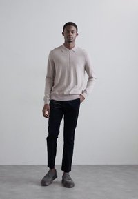 Light beige knitted sweater with a zip collar, paired with dark trousers and grey sneakers. Minimalist style with smooth textures.